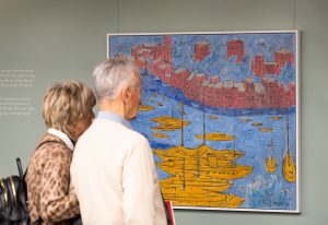 Un uomo e una donna che guardano un quadro che raffigura delle navi gialle sull'acqua e degli edifici