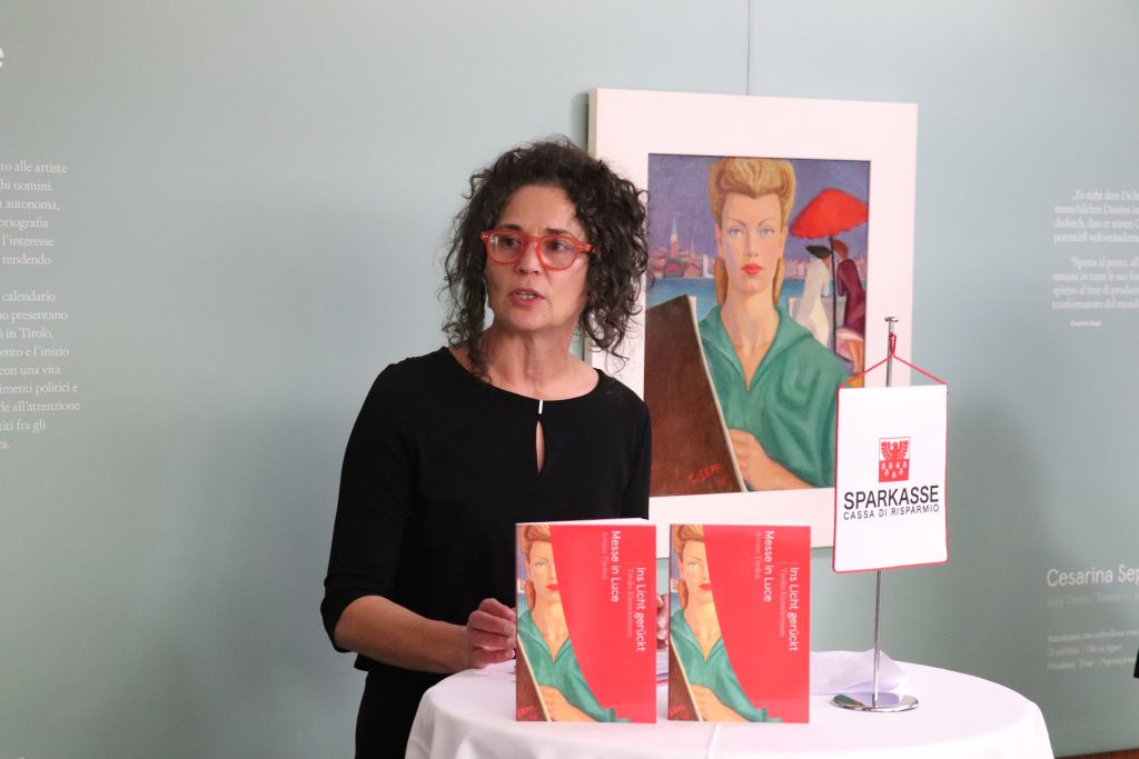 Silvia Höller con davanti 2 libri rossi e alle spalle un quadro con raffigurata una donna